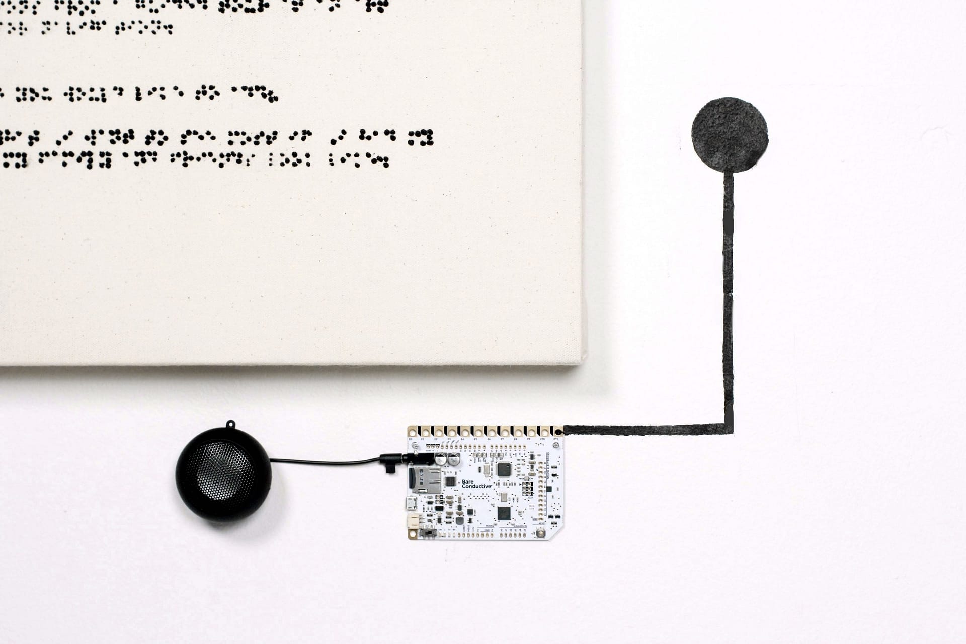 A canvas with Braille text painted with acrylic paint is mounted on a white wall. Mounted below is a small computer is attached to a speaker and conductive paint, indicating a spot to touch and activate sound.