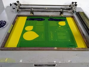 Getting the silkscreen ready to print with blobs of black ink resting above the text that will be printed.