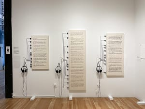 Three canvases with Braille text painted in acrylic hang on a wall. To the left of each canvas, a touch board is connected to headphones and as well as black conductive paint lines, each lining up with the question and answer in Braille.