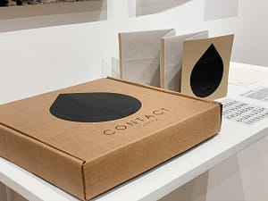 A shelf view of the contents of the CONTACT kit: a cardboard box with a black scale printed on top, an interactive accordian-style booklet, three vellum pages describing the project, a Braille booklet, and a vellum sleeve for the booklet.