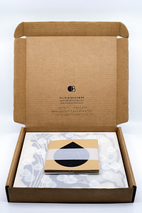 Image description: An open cardboard box with Olivia Brouwer's logo, website, and Instagram printed on the back in English text and Braille. The CONTACT artwork and booklet are displayed inside the box.