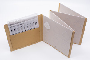 Image description: The open accordion book showing three information sheets printed on vellum tucked in a pocket. The other pages have embossed vellum slips with designated spots for the teardrop shapes to stay in. A line is embossed underneath to write out the Braille translation in English text.